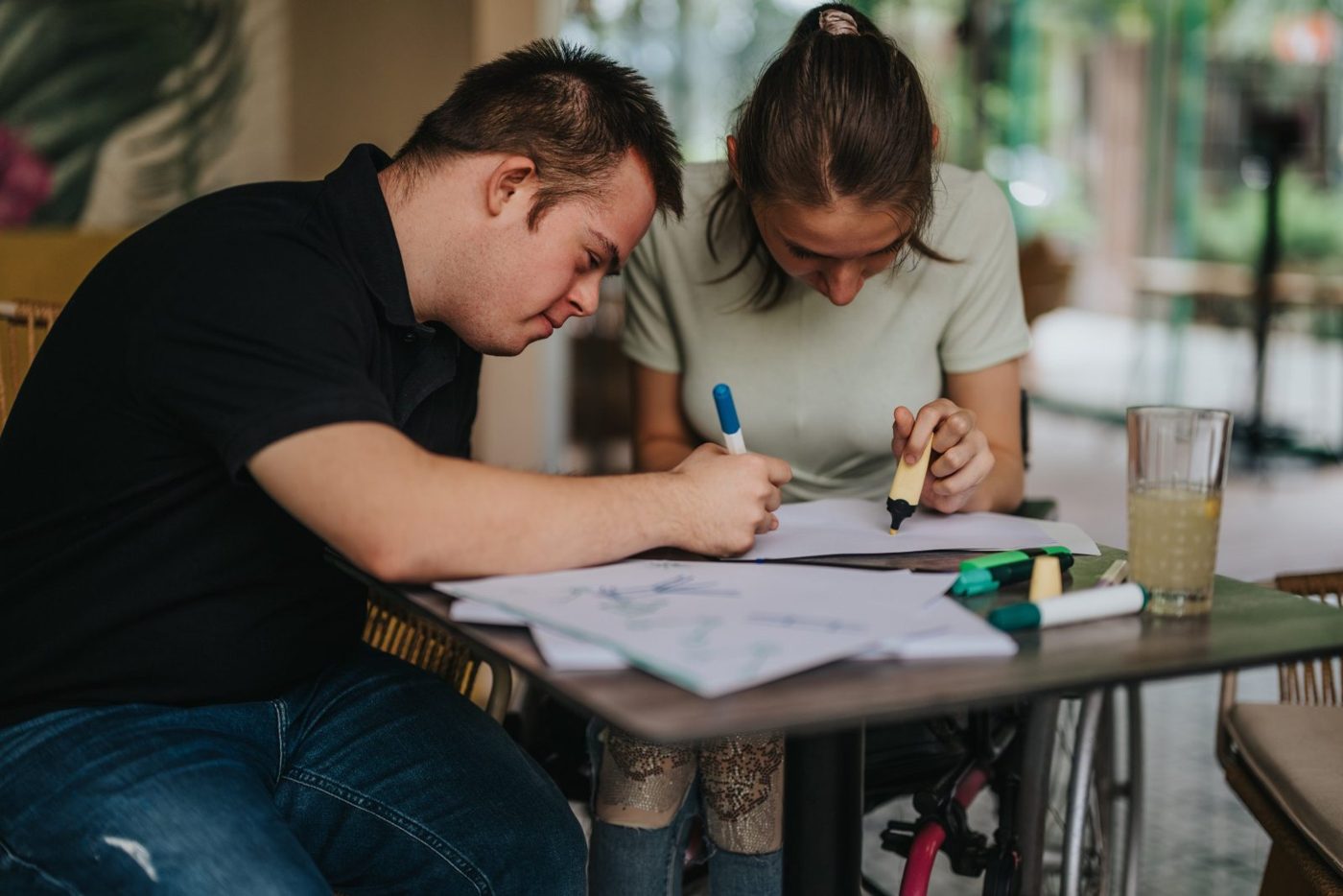 Two young adults drawing together at a café table. One of them uses a wheelchair.
